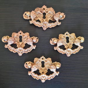 Könnte beinhalten: Vier verzierte Schubladengriffe im antiken Stil in warmem, metallischem Goldfinish. Jeder Griff hat ein Schlüsselloch und dekorative Reliefdetails mit abgerundeten Endknöpfen. Die Griffe sind auf einer dunklen Holzoberfläche angeordnet.