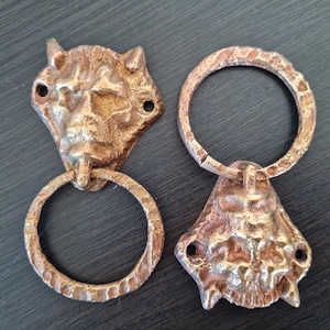 Pode incluir: Duas aldravas de porta cor bronze. Uma apresenta uma cabeça de leão com um anel, a outra um rosto estilizado com um anel. Ambas têm um aspeto texturizado e envelhecido, com orifícios de montagem.