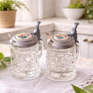 Peut inclure: Deux pots à condiments en verre transparent avec des surfaces texturées, chacun doté d'un couvercle articulé couleur étain surmonté d'un insert en céramique circulaire orné d'un motif floral coloré et d'une petite cuillère en métal.