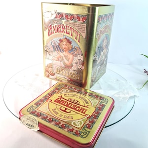 Op de afbeelding: Twee vintage Italiaanse Amaretti koektrommels. De grotere trommel is rechthoekig met een gouden afwerking en toont een vrouw met bloemen. De kleinere trommel is vierkant met een rode rand en de tekst "Gadeschi Made in Italy".