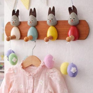 May include: A wooden wall-mounted coat rack featuring four rabbit-shaped hooks with grey heads and bodies in white, light blue, yellow, and red. Each hook holds a pair of small, felted, egg-shaped ornaments in pastel colors with white flower accents.