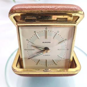 May include: A vintage travel alarm clock with a gold-tone case and a brown leatherette cover. The clock face is cream-colored with black hands and the word "EUROPA" printed at the bottom. The clock is open, revealing the face.