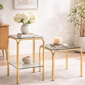 Könnte beinhalten: Ein Set aus zwei quadratischen Beistelltischen mit goldfarbenen Metallrahmen und klaren Glasplatten. Der höhere Tisch verfügt über eine untere Glasablage, während der niedrigere Tisch eine einzelne Glasplatte besitzt. Beide haben abgerundete Ecken.