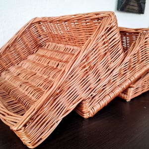 Op de afbeelding: Drie geweven rieten manden in verschillende maten. De manden hebben een warme, lichtbruine kleur en hebben een rechthoekige vorm met schuine zijkanten. Het weefpatroon is zichtbaar en voegt textuur toe. De manden zijn op een donkere ondergrond gestapeld.
