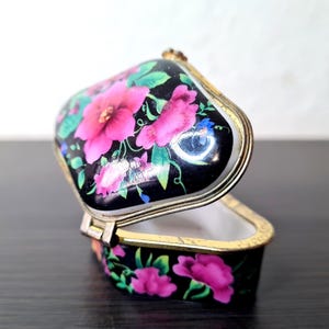 以下が含まれることがあります： 黒い背景に花柄が施された、開いた状態の小さな小物入れ。鮮やかなピンク色の花と緑の葉、金色の縁取りが特徴です。内側は白。おそらく磁器またはセラミック製です。