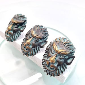 Puede incluir: Tres tiradores de armario de estilo antiguo con diseño de cara de león. Los tiradores tienen un acabado bronce con detalles turquesa y dorado, y están colocados sobre una superficie blanca. Estos tiradores decorativos son adecuados para muebles.