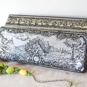 Puede incluir: Una caja metálica rectangular antigua con tapa abisagrada. La caja es de color plateado con una escena de paisaje en relieve detallada. Un collar de cuentas con cuentas amarillas y verdes está en primer plano.