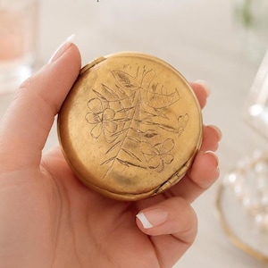 Op de afbeelding: Een rond, goudkleurig metalen doosje met een gegraveerd bloemmotief op het deksel. Het oppervlak toont een botanisch patroon met bladeren en bloemen. De tekst "A delicate keepsake for your most precious treasures." is hierboven zichtbaar.