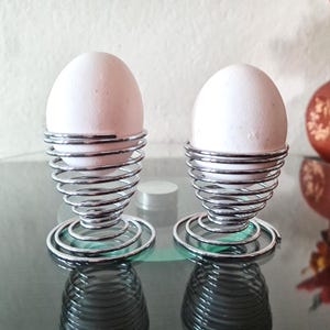 May include: Two white eggs in chrome spiral egg cups. The egg cups have a circular base and a coiled design. The background is blurred, with hints of other objects. The egg cups are made of metal.