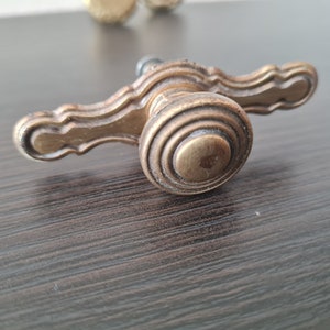 Könnte beinhalten: Antiker Schrankgriff aus Messing mit dekorativem Design. Der Griff hat einen zentralen, kreisförmigen Knopf mit konzentrischen Ringen und zwei gebogenen Griffen. Die Hardware hat eine warme, gealterte Messingfarbe, die einen Vintage- oder Retro-Stil suggeriert.