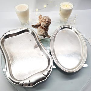 Könnte beinhalten: Zwei silberfarbene Serviertabletts, eines rechteckig mit gewelltem Rand und eines oval mit glattem Rand, platziert neben einer kleinen beigen Cherub-Figur und zwei mit weißem Wachs gefüllten Glas-Kerzen, die von einer Lichterkette beleuchtet werden.