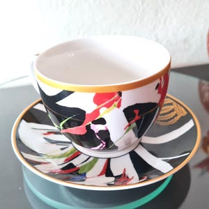 Puede incluir: Un juego de taza y platillo blancos con un vibrante diseño floral en rojo, negro y blanco. La taza tiene un borde dorado y el platillo presenta un patrón a juego. Una elegante adición a cualquier juego de té.