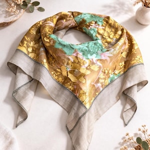 May include: A square scarf with a floral pattern in shades of gold, brown, and teal. The scarf has a light gray border with black stitching. The scarf is folded in a triangle shape.