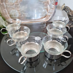 Può includere: Set di sei tazze da tè in vetro trasparente con manici e basi in metallo argentato. Le tazze sono disposte su un vassoio di vetro a specchio, con un vassoio d'argento e una statua decorativa di gallo sullo sfondo.