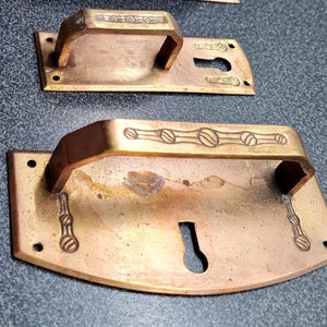 Könnte beinhalten: Zwei antike Türgriffe aus Bronze. Die Griffe haben eine rechteckige Rückplatte mit Schlüsselloch und einen gebogenen Griff. Die Griffe sind mit einem eingravierten Design aus Kreisen und Linien verziert.