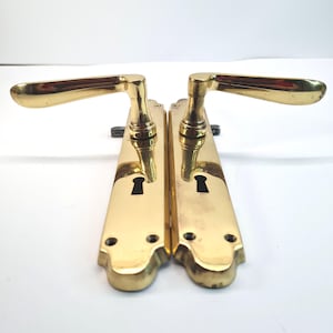 以下が含まれることがあります： 真鍮製のドアノブ2個セット。各ノブは、鍵穴と湾曲したレバーを備えた長方形のバックプレートを備えています。ノブは光沢のある金色で、ヴィンテージのようです。