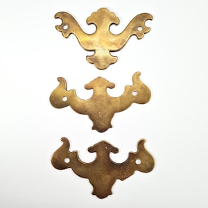 Puede incluir: Tres tiradores de cajón antiguos de latón con un diseño decorativo y simétrico. Cada tirador presenta un elemento central con brazos curvos que se extienden hacia afuera y pequeños orificios para el montaje. Los tiradores tienen una pátina cálida y envejecida.