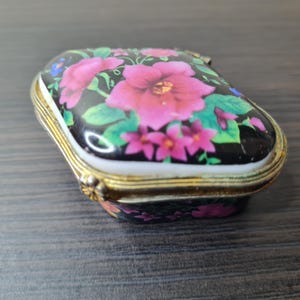 May include: A small, rectangular trinket box with a hinged lid. The lid is decorated with a floral pattern featuring pink flowers and green leaves on a black background. The box has gold-colored metal trim.