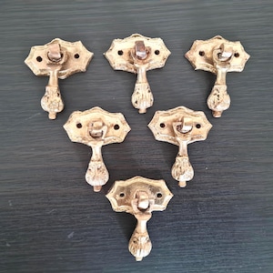 May include: Six antique brass drawer pulls on a dark surface. Each pull has an ornate backplate with a central handle and a detailed, teardrop-shaped pull. The brass has a slightly aged, vintage appearance, showing signs of age and use.