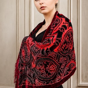 May include: A rectangular velvet shawl featuring a deep red and black color palette with an intricate, repeating pattern of ornate, shield-like motifs and decorative scrollwork. The fabric has a soft, textured finish with fringed edges.