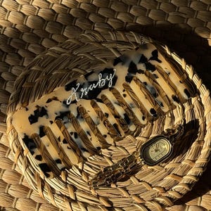 May include: A tortoiseshell-patterned comb with the word "Kraby" in cursive script. The comb rests on a woven basket, accompanied by a gold wristwatch. The comb's design features a mix of tan and black spots.