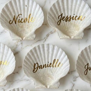 May include: A collection of white scallop shells used as decorative dishes, each featuring a name written in metallic gold cursive script. The visible names include Nicole, Jessica, and Danielle, centred on the interior of the ribbed shells.