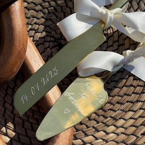 May include: Gold-coloured cake knife and server set with white ribbon bows. The knife is engraved with the date "14.07.2025", and the server says "here's to forever" with a heart symbol. The set is on a woven surface.