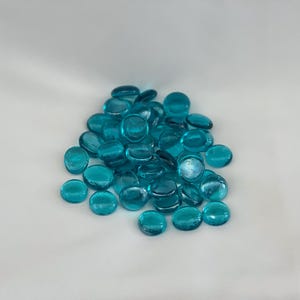 May include: A pile of turquoise glass pebbles. The smooth, rounded stones are a vibrant teal color and appear translucent. They are scattered on a white surface, creating a visually appealing contrast.