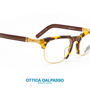 May include: A pair of tortoiseshell eyeglasses with gold metal frames and burgundy acetate temples. The temples have a gold stripe running along the top and bottom. The eyeglasses are on a white background.