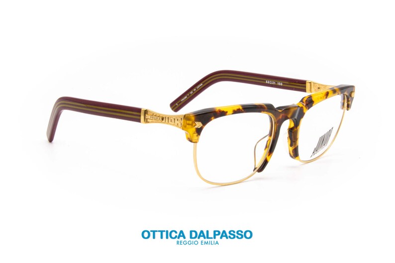May include: A pair of tortoiseshell eyeglasses with gold metal frames and burgundy acetate temples. The temples have a gold stripe running along the top and bottom. The eyeglasses are on a white background.