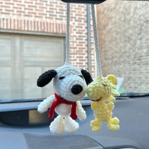 Könnte beinhalten: Zwei gehäkelte Stofftiere hängen am Rückspiegel eines Autos. Das weiße und schwarze Spielzeug ist Snoopy, der einen roten Schal trägt. Das gelbe Spielzeug ist Woodstock. Das Armaturenbrett des Autos ist im Hintergrund zu sehen.
