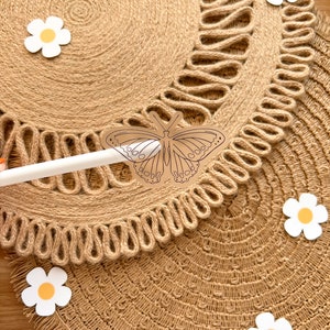 May include: Close-up of two round, woven jute placemats with a decorative edge. A butterfly-shaped cutout is held by a white stylus. White daisy cutouts with yellow centres are scattered around the placemats.