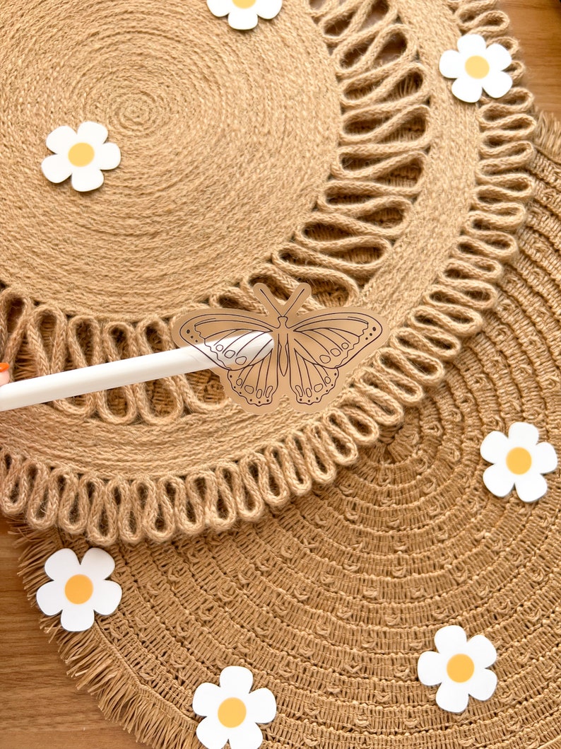 May include: Close-up of two round, woven jute placemats with a decorative edge. A butterfly-shaped cutout is held by a white stylus. White daisy cutouts with yellow centres are scattered around the placemats.