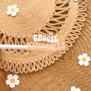 May include: A sticker with the text "Choose Kindness" in a retro font with pink and orange colors and daisy accents on a brown woven placemat.