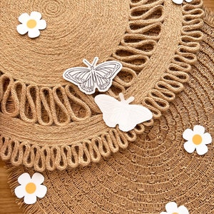 May include: Two round, woven jute placemats with a decorative edge. White butterfly and daisy cutouts are scattered across the surface. The placemats are a natural tan colour, with a fringed edge.