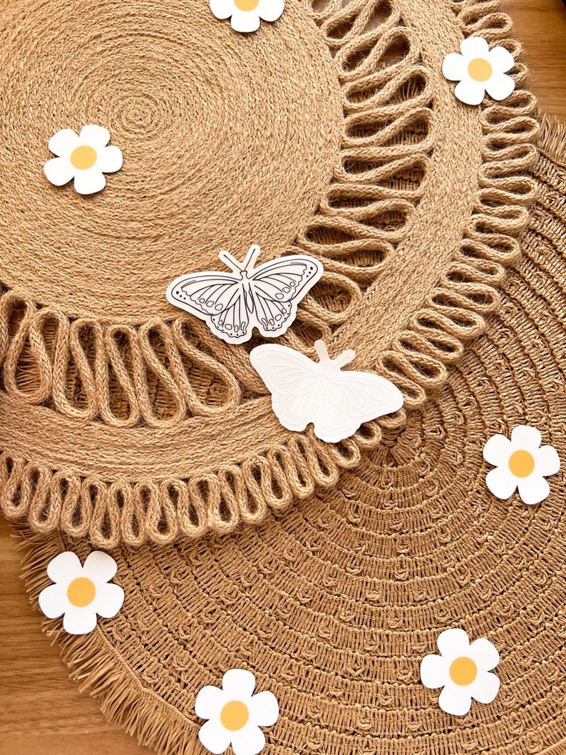 May include: Two round, woven jute placemats with a decorative edge. White butterfly and daisy cutouts are scattered across the surface. The placemats are a natural tan colour, with a fringed edge.
