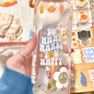 May include: Clear glass water bottle with a bamboo lid and a tan strap. The bottle is decorated with stickers, including autumn leaves, a butterfly, and the phrase "Do What Makes You Happy."