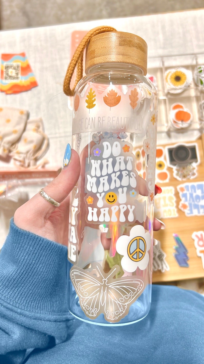 May include: Clear glass water bottle with a bamboo lid and a tan strap. The bottle is decorated with stickers, including autumn leaves, a butterfly, and the phrase "Do What Makes You Happy."