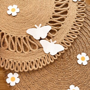 May include: Two round jute placemats with a decorative edge. White butterfly and daisy cutouts are scattered across the surface. The placemats are a natural tan colour, with a fringed edge.