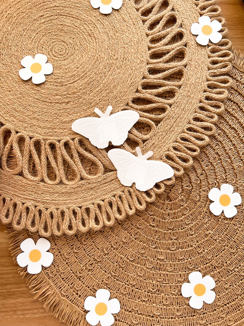 May include: Two round jute placemats with a decorative edge. White butterfly and daisy cutouts are scattered across the surface. The placemats are a natural tan colour, with a fringed edge.