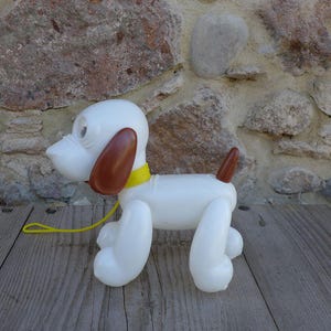 Può includere: Un cane giocattolo vintage in plastica bianca con orecchie e coda marroni, e un collare e guinzaglio gialli. Il cane è posato su una superficie di legno contro un muro di pietra.