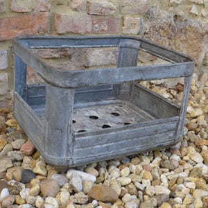 Könnte beinhalten: Ein verwitterter, quadratischer Metallbehälter mit perforiertem Boden und offenen Seiten. Der Behälter besteht aus verzinktem Stahl und steht auf einem Bett aus kleinen, bunten Steinen. Der Hintergrund zeigt eine Backsteinmauer.