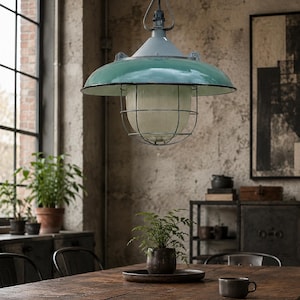 Op de afbeelding: Een hangende pendellamp met een brede, glanzend groenblauwe metalen kap, een centrale geribbelde glazen diffuser en een beschermende donkere metalen draadkooi. De lamp is opgehangen aan een ketting en een metalen beugel.