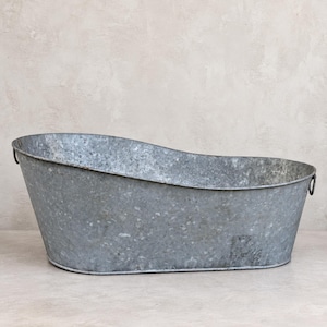 Könnte beinhalten: Eine Wanne aus verzinktem Metall mit einer verwitterten, gesprenkelten grauen Oberfläche. Das Becken hat eine längliche, ovale Form mit einem leicht erhöhten hinteren Rand und kleinen, runden Metallgriffen an jeder Seite des Randes zum Tragen.