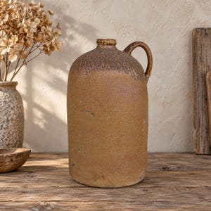 Pode incluir: Um jarro cilíndrico de grés com um esmalte texturizado e manchado em tons de castanho e bege. O recipiente apresenta um gargalo estreito, uma asa curva na lateral e um acabamento mais escuro e rugoso em torno do ombro e da borda superior.