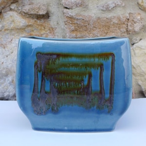 May include: A rectangular ceramic vase with a smooth, glossy blue glaze. The centre of the vase features a decorative, abstract drip pattern in shades of dark brown and olive green that runs vertically down the surface of the blue ceramic body.