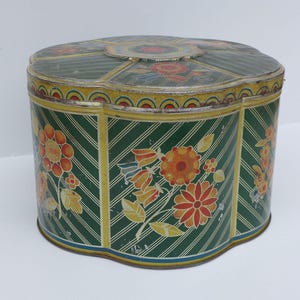 May include: A clover-shaped metal storage tin featuring a dark green background with diagonal white stripes and a floral pattern in shades of orange, red, and yellow. The lid has a matching floral design and a yellow border with a red and blue arc pattern.