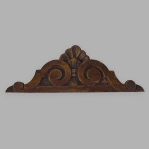Può includere: Un frontone architettonico in legno marrone scuro caratterizzato da un motivo centrale intagliato a ventaglio affiancato da motivi a volute simmetrici. L'elemento decorativo presenta una base piatta e un bordo superiore curvo con rilievi.