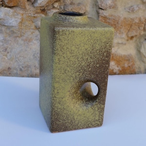 Puede incluir: Un jarrón rectangular de cerámica con un acabado texturizado y moteado en tonos verde oliva y marrón oscuro. El jarrón presenta un recorte circular en el lado inferior derecho y una pequeña abertura circular en la parte superior.