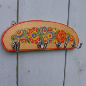Puede incluir: Perchero de pared de madera con diseño floral. El perchero tiene una base de madera clara con un borde rojo y cuatro ganchos de metal. El patrón floral incluye flores rojas, amarillas, azules y verdes.